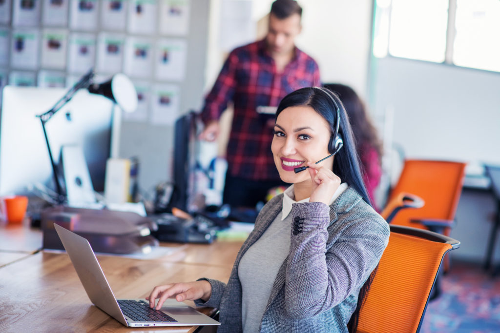 Beautiful business people in headsets are using computers and smiling while working in office. Girl is looking at camera